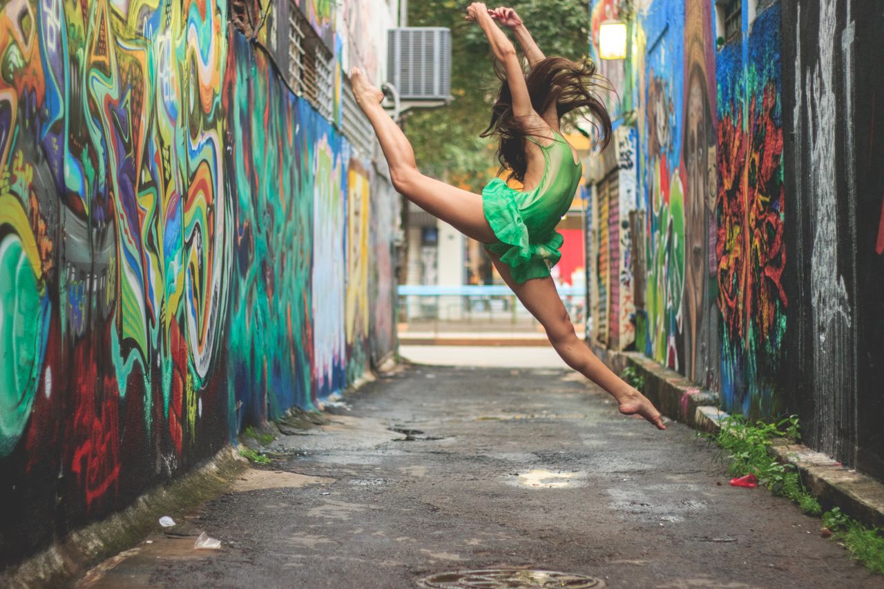 A woman performs an energetic dance leap in a colorful graffiti-filled alleyway, showcasing vibrant urban expression.